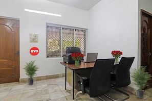 A small office with a desk, two chairs, a laptop, a printer, and a window with a metal gate. There are two potted plants and a door with a sign.