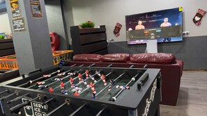 A room with a foosball table, a television mounted on the wall, and a seating area with a red couch. - hotel Kapriz Karakol (Karakol)