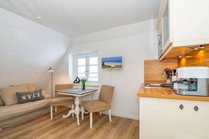 A compact living space with a kitchenette, a dining area with a table and chairs, a sofa with cushions, and a window with a view. - Holiday apartment in Sylt with swimming pool (Sylt)