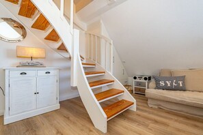 A staircase with wooden steps and white railings, a white cabinet with drawers, a round mirror, a lamp, a beige sofa with a cushion, and a small table with speakers. - Holiday apartment in Sylt with swimming pool (Sylt)