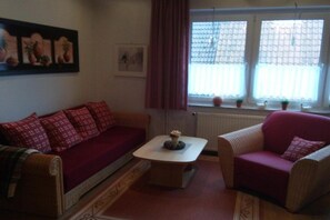 A living room with a sofa, armchair, and a coffee table. There is a window with curtains and a radiator. - Holiday apartment with 1 bedroom (Cuxhaven - Duhnen)