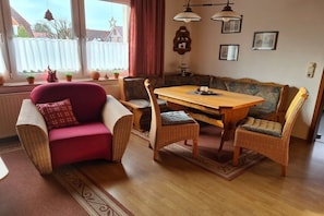 A cozy living room with a red armchair, a wooden dining table, wicker chairs, a patterned sofa, and a view of a white fence outside. - Holiday apartment with 1 bedroom (Cuxhaven - Duhnen)