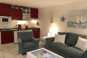 Un espacio habitable compacto con kitchenette, un sofá gris y un cuadro de barcos de vela en la pared.