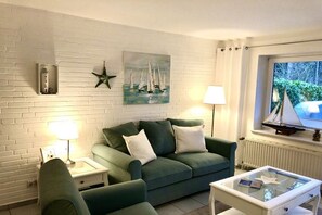 A room with a green sofa, a glass coffee table, a lamp, a painting of sailboats, a starfish decoration, and a model sailboat on the windowsill. - Holiday apartment with 1 bedroom (Hohwacht)