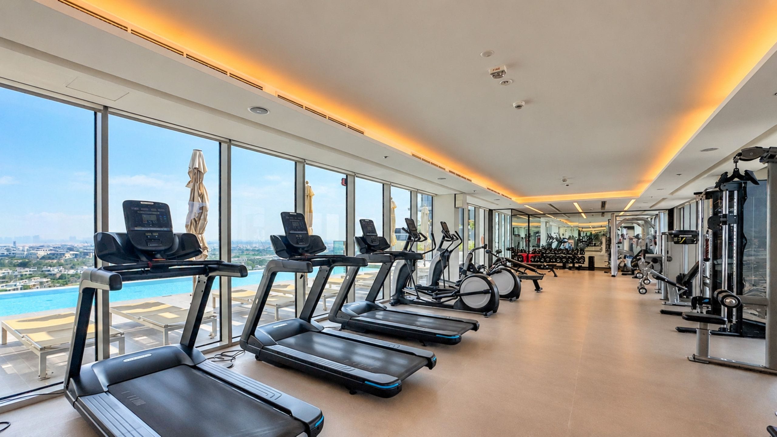 Ein Fitnessstudio mit Laufbändern, Ergometer und Krafttrainingsgeräten, großen Fenstern mit Blick auf die Stadt und einer Decke mit Einbuchtungslichtern.