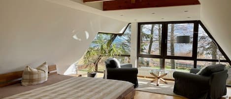 A bedroom with a large bed, a chair, and a small table. There is a window with a view of trees and a fence.