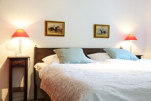 Een netjes opgemaakt bed met een kleurenschema in het wit en lichtblauw, twee nachtkastjes met lampen, en twee ingelijste foto's aan de muur.