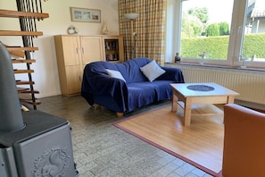 A cozy living room with a blue sofa, a wooden table, a bookshelf, and a window with a view of greenery. - Holiday home in Langholz with swimming pool (Langholz)