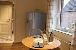 A small dining area with a round table, a chair, a refrigerator, a window with curtains, and a radiator. - Holiday home "Mittenmang" (Schleswig)