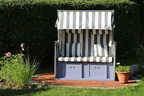 A garden seating area with a striped awning, a bench with cushions, and a potted plant. - Holiday home "Mittenmang" (Schleswig)