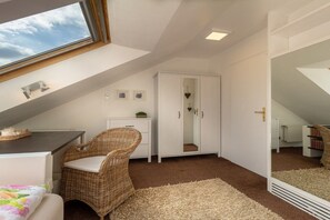 A room with a sloped ceiling, a large window, a wicker chair, a white wardrobe, and a carpeted floor. - Exclusive holiday apartment with views of the Harz Mountains (Goslar)
