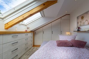 Quarto com claraboia, cama com almofadas cor-de-rosa, penteadeira e luminária fixada na parede.