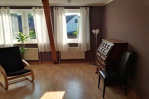 A room with a wooden floor, two black chairs, a radiator, a window with white curtains, and a small plant. - Holiday apartment with 2 bedrooms (Panker\/ Gadendorf)