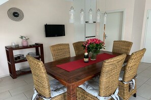Ein Essbereich mit einem Holztisch, Korbstühlen, einer roten Tischmatte, einer Flasche und einer Vase mit Blumen.