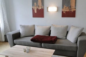 A grey sofa with striped cushions and a red throw blanket, a wooden coffee table with glasses, and two wall-mounted light fixtures. - Holiday apartment with 2 bedrooms (Kappeln Stadt)