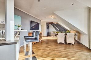 Een ruime woonkamer met houten vloer, een eettafel met stoelen, een bank en een trap naar een hoger niveau.
