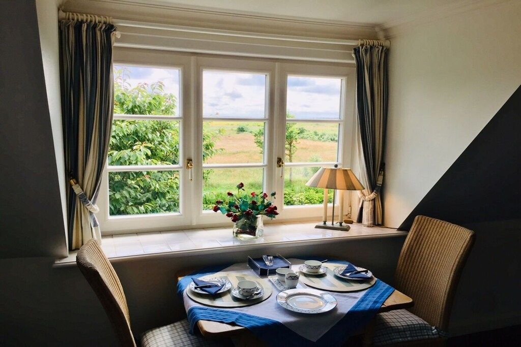 A dining area with a table set for two, a window with curtains, and a view of greenery outside.