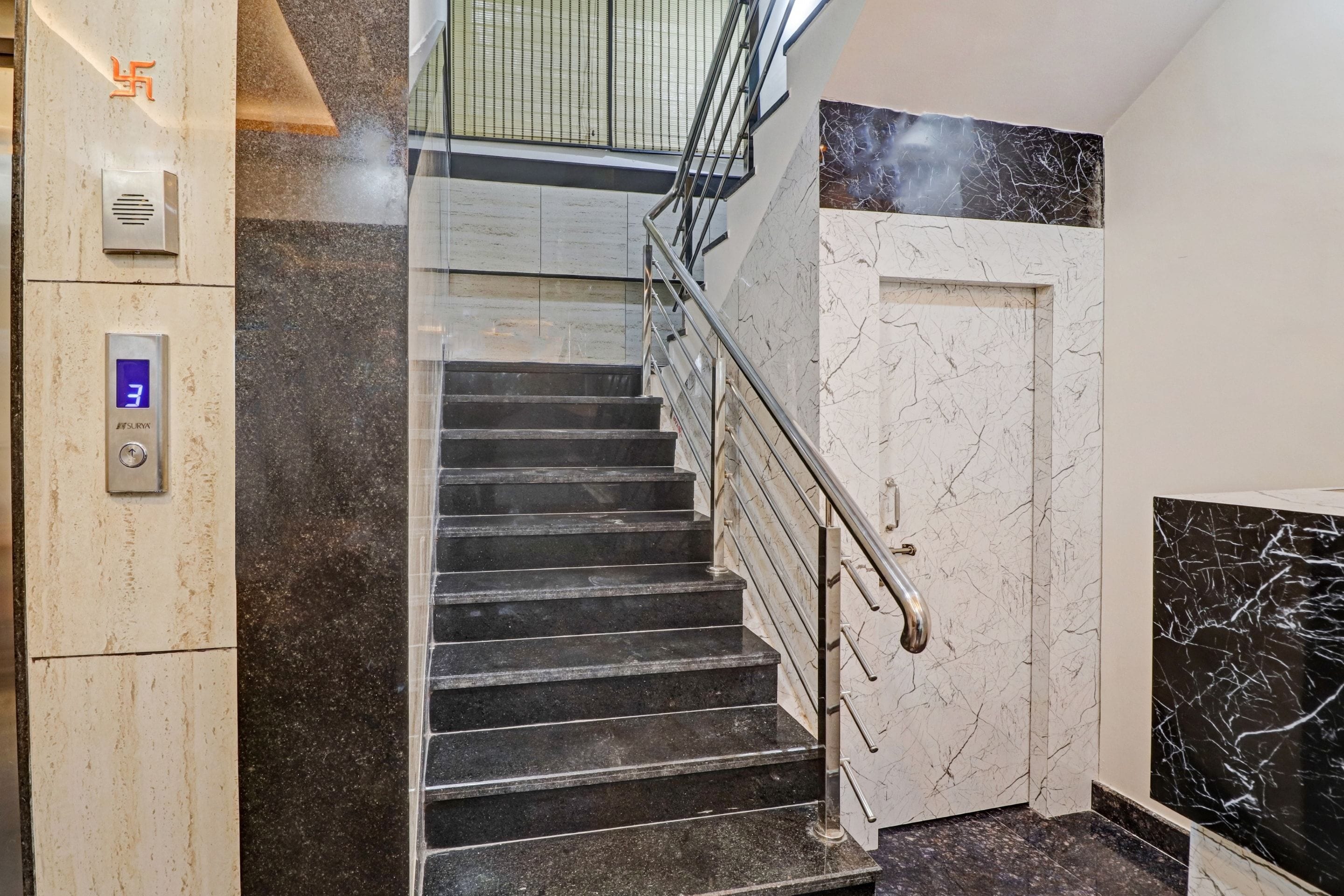 Un escalier avec une rampe, une porte avec un revêtement en marbre et un panneau de commande d’ascenseur fixé au mur.