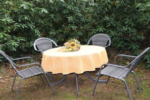 A round table with a yellow tablecloth, surrounded by four grey wicker chairs, set outdoors with a backdrop of dense green foliage. - Holiday apartment in Schneverdingen with Wi-Fi (Schneverdingen)