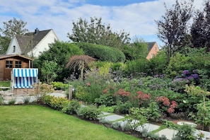 A garden with a variety of plants and flowers, a wooden structure with a striped awning, and a house in the background. - Stylish 3-room holiday apartment "City Oasis" (Westerland)