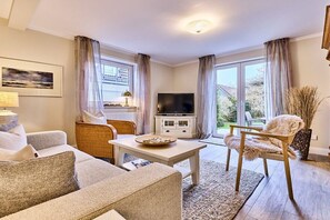 Ein Wohnzimmer mit einer Couch, Sesseln, einem Couchtisch, einem Fernseher und einem großen Fenster mit Vorhängen.