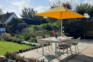 An outdoor patio with a table and chairs, a yellow umbrella, and a garden with various plants and a wooden fence. - Stylish 3-room holiday apartment "City Oasis" (Westerland)