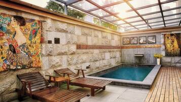 An indoor swimming pool area with wooden loungers, a stone wall with artwork, and a glass ceiling.