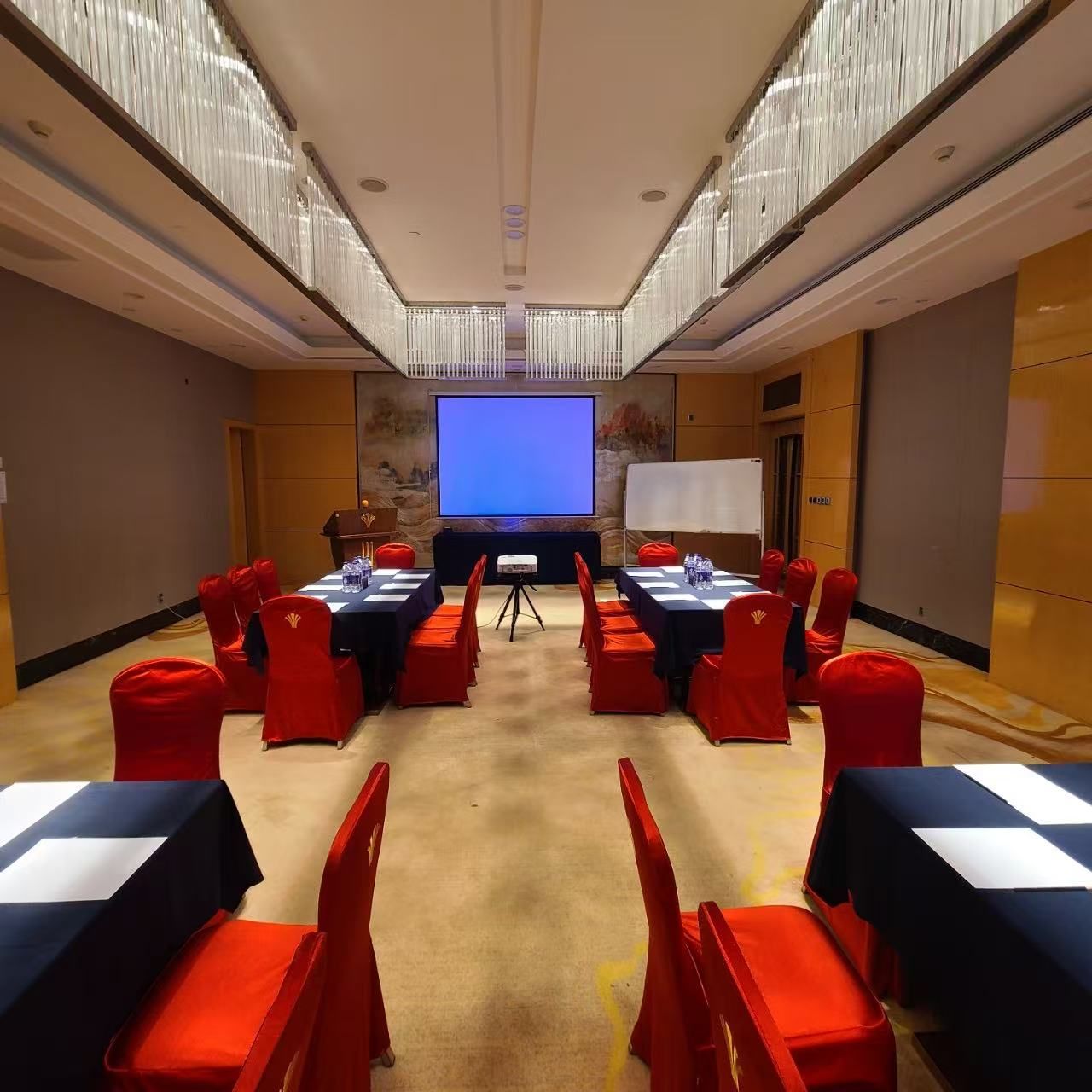 Un salón de conferencias con una pantalla grande, una pizarra y sillas rojas alrededor de una mesa.