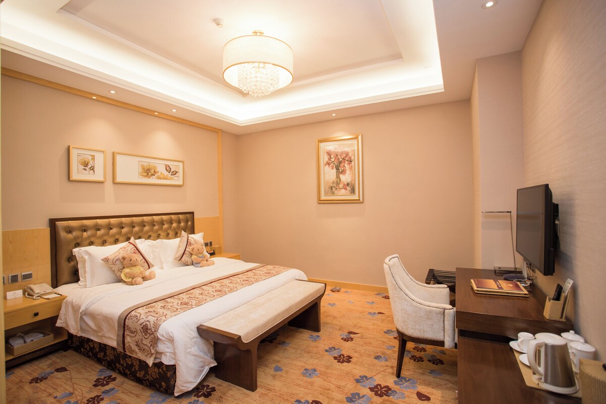 Una habitación de hotel con una cama grande, mesitas de noche, un escritorio con silla, televisión y obras de arte en las paredes.