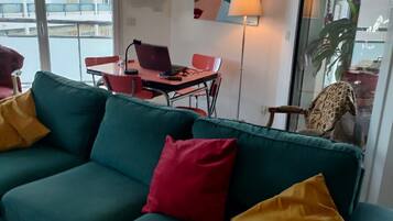 Un salon avec un canapé vert, des coussins rouges et jaunes, un ordinateur portable posé sur une petite table, et un tableau accroché au mur.