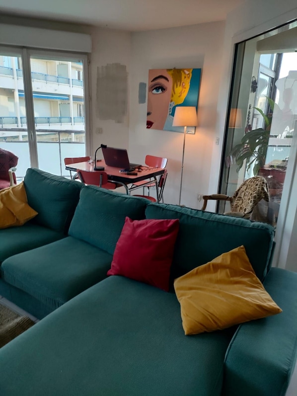 A living room with a green sofa, red and yellow cushions, a laptop on a small table, and a painting on the wall.