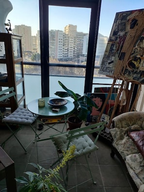 A room with a view of a cityscape, a round table with a potted plant, a sofa, and a wall with a collage of images and text.