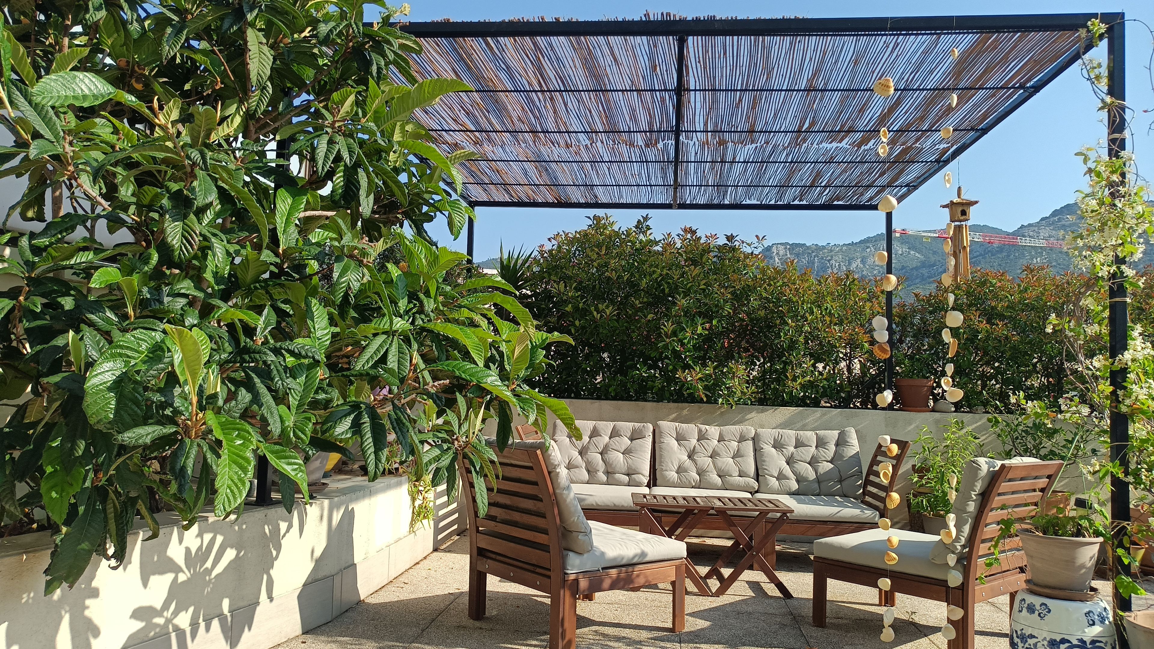 Un área de estar al aire libre con sofá, sillas y una mesa, rodeada de plantas en macetas y una valla decorativa.