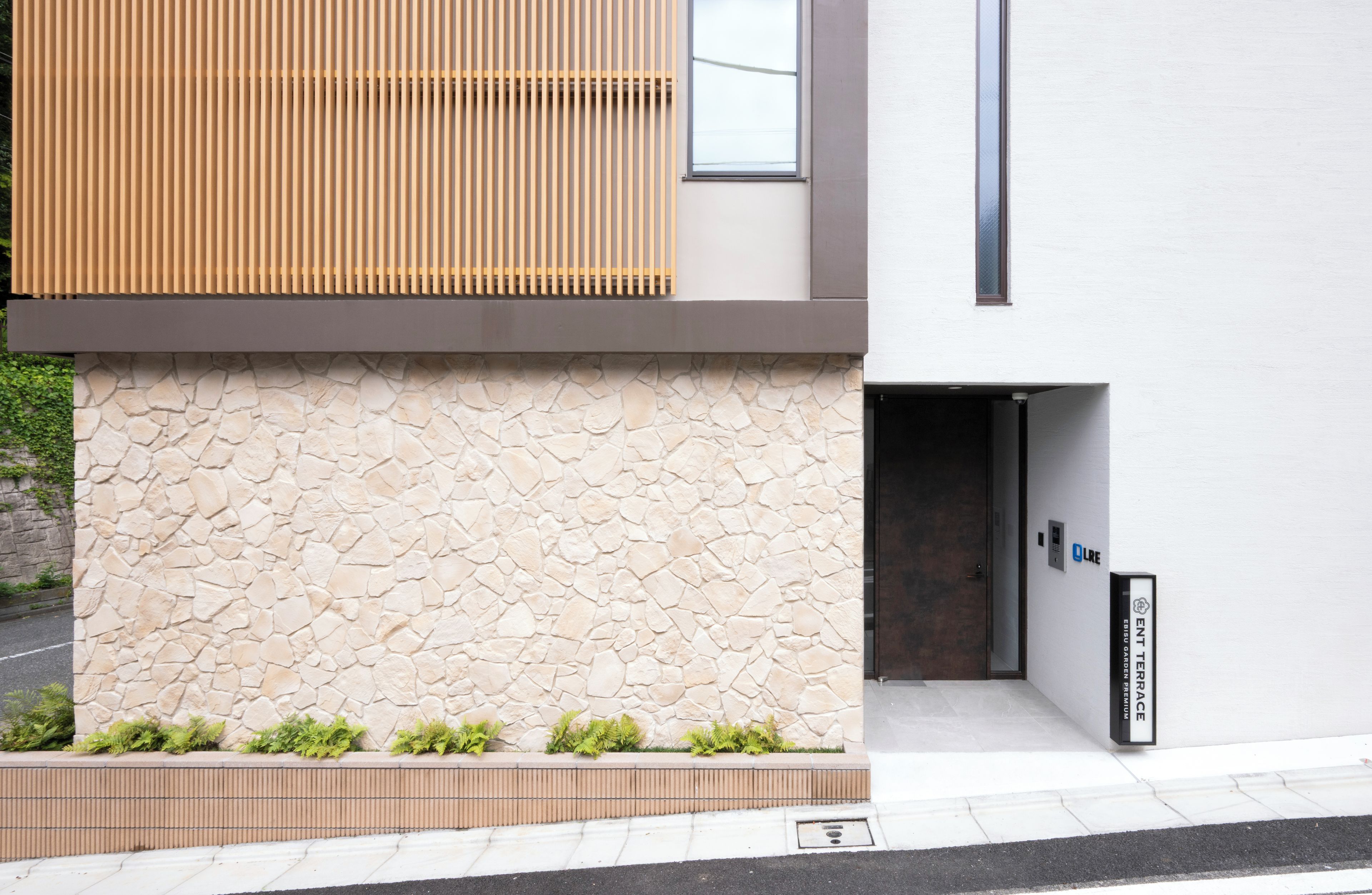 A modern building facade with a stone wall, a wooden slatted structure, and a door with a sign.