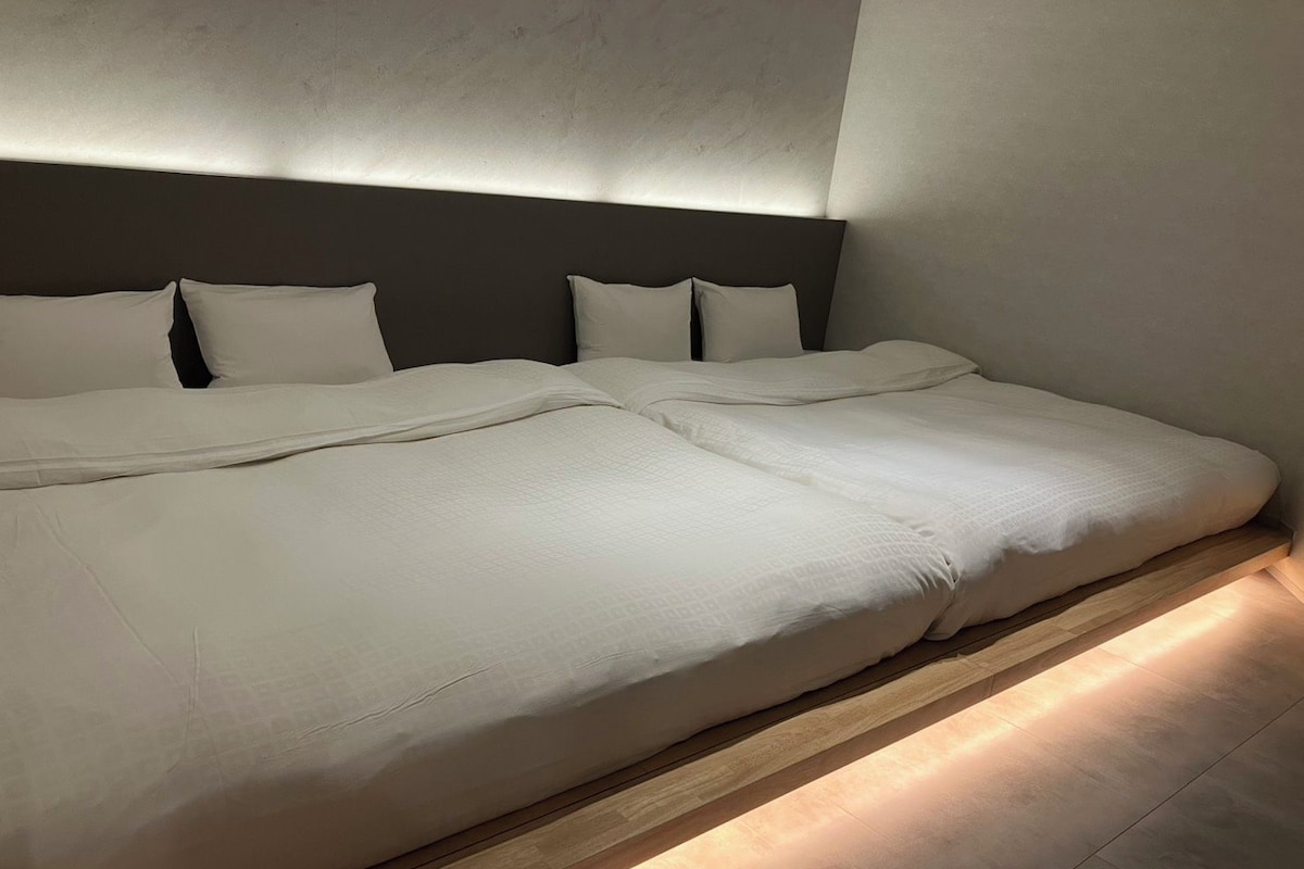 Una cama grande con cabecera y varias almohadas, suelo de madera y pared con textura.