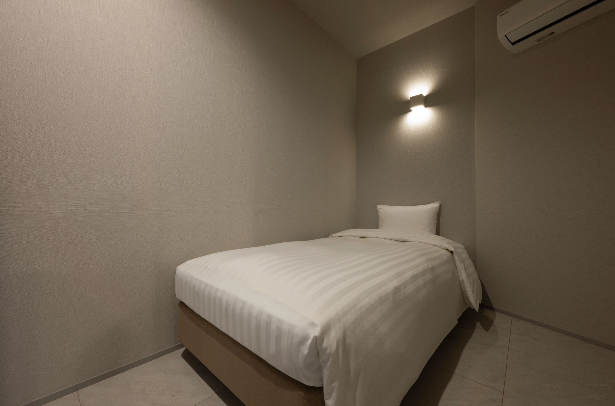 Una habitación pequeña con una cama individual, pared sencilla, aire acondicionado instalado en el techo y una lámpara fijada a la pared.