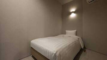 Una habitación pequeña con una cama individual, pared sencilla, aire acondicionado instalado en el techo y una lámpara fijada a la pared.