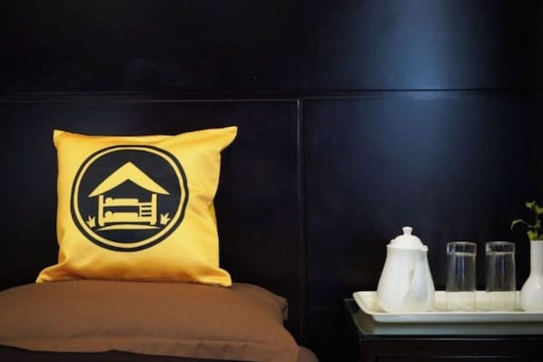 A bed with a yellow pillow featuring a black and white logo, a brown mattress, and a bedside table with a white pitcher, two glasses, and a vase with a plant.
