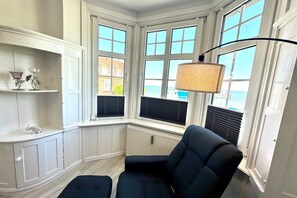 Ein Eckzimmer mit Erkerfenster, einem schwarzen Sessel, einem Regal mit Dekoration und einer Heizung.