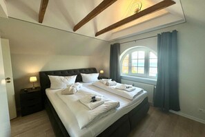 A double bed with white linens and towels, a nightstand with a lamp, a window with curtains, and a wooden ceiling beam. - House with sea view - Apt. 4 (Wyk auf Föhr)