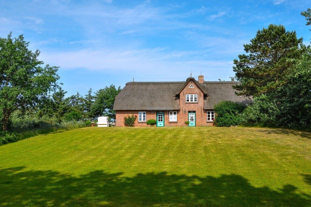 Une maison à un étage, construite en briques et coiffée d’un toit de chaume, entourée d’une pelouse bien entretenue et d’arbres.