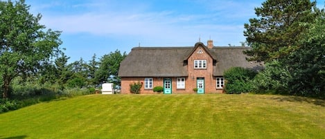 Une maison à un étage, construite en briques et coiffée d’un toit de chaume, entourée d’une pelouse bien entretenue et d’arbres.