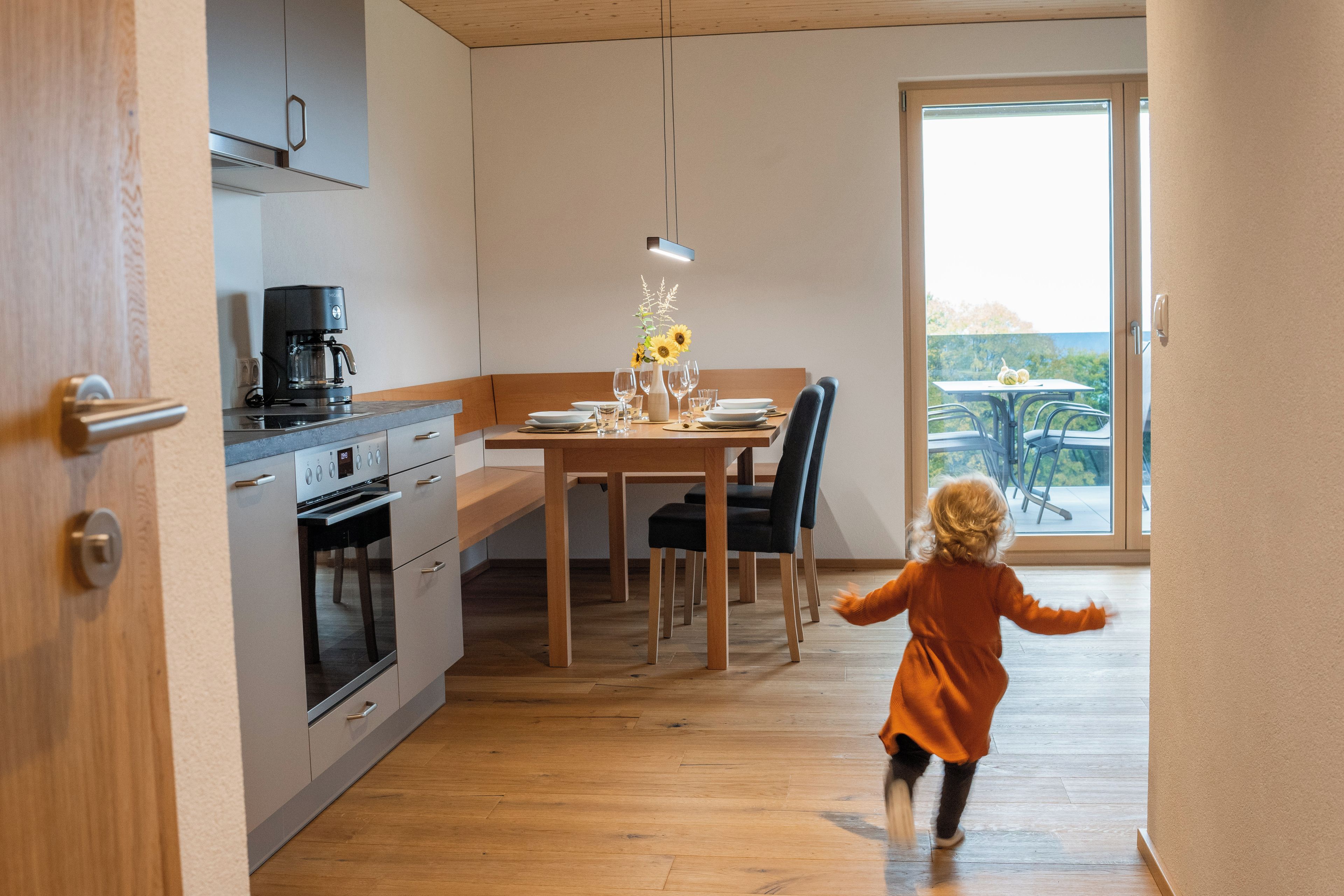 Une cuisine moderne avec un coin repas, une machine à café et un enfant qui court vers un balcon.