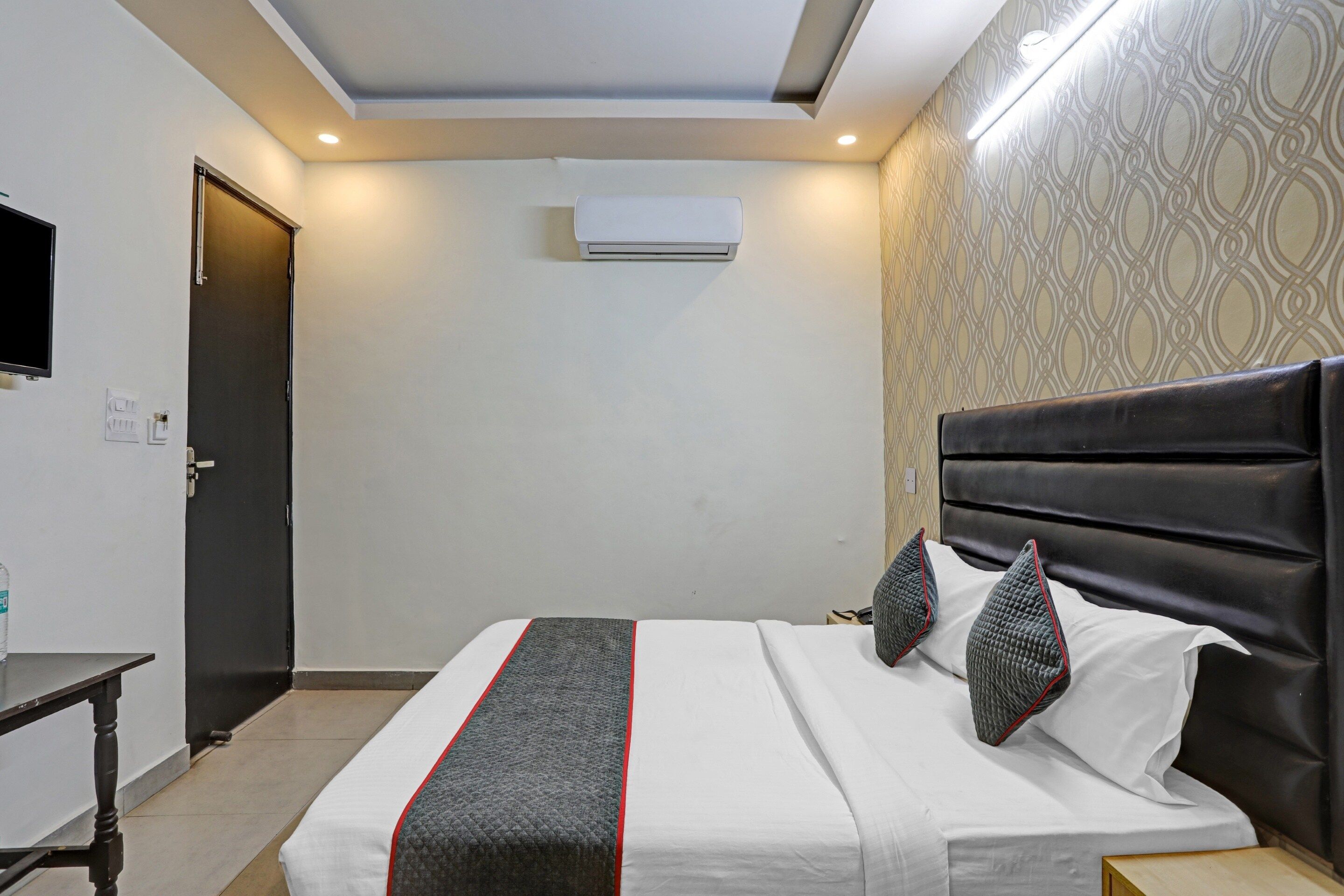 A hotel room with a bed, a bedside table, a wardrobe, a flat-screen TV, and an air conditioning unit.