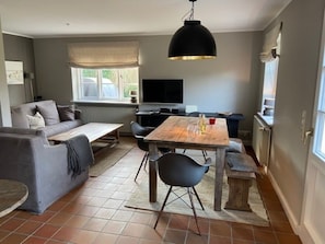 A living room with a sofa, a coffee table, and a dining table with chairs. There is a television on the wall, a window with a view, and a hanging light fixture. - 8a in the Building on Neuer Weg (Wrixum)