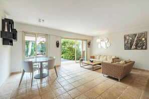 A spacious living area with a dining table, sofa, and a large window offering a view of greenery. - Family Apartment With Pool & Garden 5 P - Cannes (Cannes)