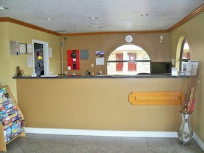 A reception area with a counter, a fire alarm, a calendar, and a window with a view of a building. - Scot Tish Inns And Suites (Beaumont)