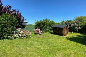 A wooden barrel structure in a grassy area with flowering bushes and trees, a clear sky, and a body of water in the background. - Holiday home in Waabs with swimming pool (Waabs)
