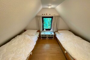 A small, triangular attic bedroom with two single beds, a small table, and a window with curtains. - Holiday home in Waabs with swimming pool (Waabs)
