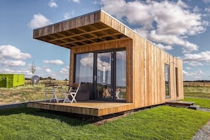 A modern wooden cabin with a glass door entrance, a deck with seating, and a clear sky in the background. - Holiday home with 2 bedrooms (Schwedeneck)